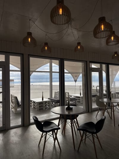 The image displays the interior of a dining establishment located in Yantarny, Russia, with a panoramic view of a sandy beach and the sea. The indoor area features a smooth, light-colored floor and a white, paneled ceiling from which several industrial-style pendant lights with exposed filament bulbs and wooden slatted shades are suspended by black wiring.

In the foreground and midground, two circular light-colored tables with splayed wooden legs are visible. Each table is accompanied by three black Eames-style chairs, also with splayed wooden legs. The central table holds a dark bottle or vase containing dried plant material.

Beyond a wall of multi-paneled glass, an outdoor wooden deck is visible. This deck is furnished with multiple seating areas, each comprising low tables and benches constructed from white-painted wooden pallets. Some benches are fitted with dark cushions. Overhead, white tensile fabric canopies provide partial cover for the outdoor seating.

The deck transitions directly to a wide, light-colored sandy beach. The sea, appearing grey under an overcast sky, stretches into the distance. Three individuals are observed walking on the beach in the mid-distance; two are together towards the left, and one is further right. A wooden structure is present on the far left of the beach, and another structure is visible near the waterline on the right. No individuals are present within the depicted indoor space.