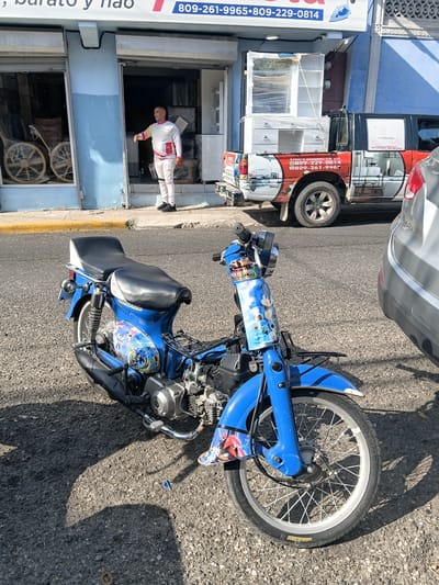 In the city of Puerto Plata, Dominican Republic, a bright blue moped with Sonic the Hedgehog decals is parked on an asphalt street in front of a storefront. The moped is the primary subject, with its engine, wheels, and seat clearly visible. A man in a white shirt and light-colored pants stands near the entrance of the store, gesturing with his left hand. Behind him, the store appears to be a furniture shop, with large furniture items, some wrapped in plastic, visible inside and on a red and white pickup truck parked in front of the store. The truck is branded with "MUEBLERÍA ACOSTA" and phone numbers. The building has a blue facade, and the street is lined with a yellow curb. The scene captures a moment of commercial activity on a sunny day.