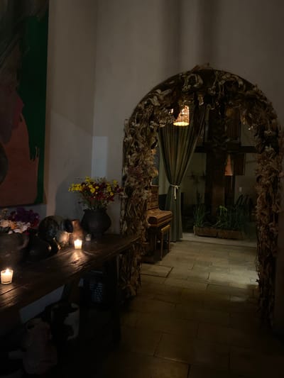 This image depicts an interior space in Oaxaca City, Mexico, likely a restaurant or a decorative corner of a building. The most prominent feature is a large archway adorned with dried plant matter, creating a rustic and naturalistic frame. Through the archway, a dimly lit passage is visible, revealing a stone-paved floor and some plants growing in containers. The space beyond the arch has a vaguely domestic feel, with glimpses of what might be furniture and other objects. In the foreground, along the wall, is a wooden surface displaying several earthen vases and a few lit candles, adding a touch of warmth and ambiance. The color palette is dark and muted, with highlights provided by the candles and the yellow and red hues of the flowers in one of the vases. A painting on the wall is partially visible in the lower left corner. Overall, the scene suggests a carefully curated, atmospheric space that emphasizes natural elements and a sense of rustic elegance.
