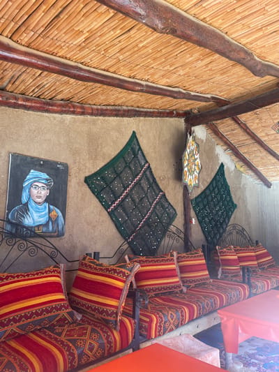 A corridor features a wall crafted from natural materials like bamboo. The floor is concrete, adorned with handwoven rugs and a painted portrait of a man wearing glasses and a blue turban. Cushioned seating with red and yellow striped pillows line the opposite side, their ornate iron frames visible. A decorative star-shaped tile sits near the wall.