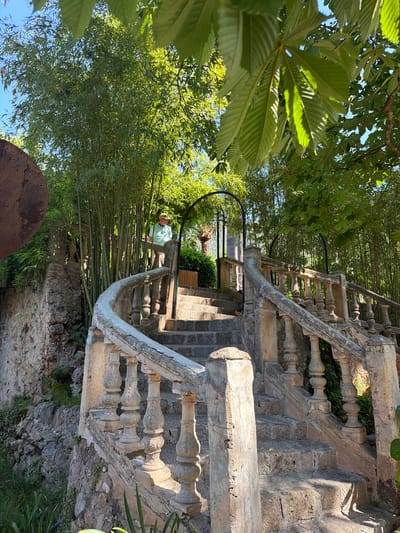 The image depicts a stone staircase with an ornate balustrade in a garden setting, likely in Bunyola, Spain. The stairs are aged and worn, leading upwards through lush vegetation that includes trees, bamboo, and other plants. Sunlight filters through the foliage, creating dappled patterns on the steps and balustrade. A person wearing a light-colored shirt can be seen partially obscured by the vegetation. In the foreground, a large, dark, possibly rusted object is partially visible. The scene conveys a sense of historical architecture integrated into a natural environment.