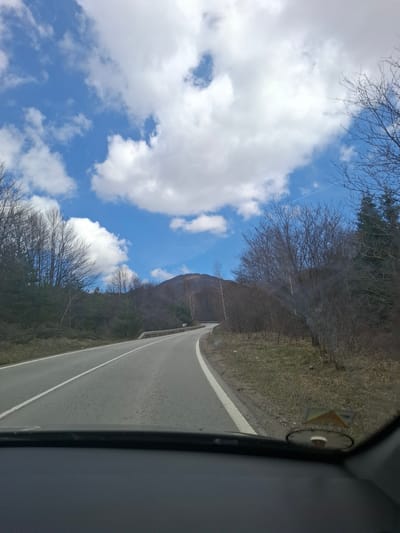 The image depicts a view from inside a vehicle, likely a car, driving on a road in what appears to be a mountainous area near Gintsi, Bulgaria. The road is paved with a white line marking the lanes. To the side of the road, there's sparse vegetation and bare trees, suggesting either late autumn or early spring. In the distance, a mountain is visible under a sky with blue patches and scattered white clouds.