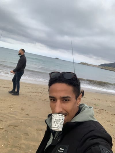 A selfie-style photo captures two men on a cloudy day at a beach in M'diq, Morocco.

In the foreground, a man with dark hair and a light beard/stubble is seen from the chest up, taking the photo. He wears sunglasses pushed onto his head and is dressed in a black jacket over a light-colored hooded sweatshirt. He holds a white disposable coffee cup to his mouth, with a slight smile on his face. Visible text on the cup includes "COFFEE", "ESPRESSO", "Instant Coffee", "CAFE CUP", and "CAPPUCCINO". On his jacket, a patch with the text "-18-" and "SHUGHUYTR" is visible.

Further back on the sandy beach, another man stands facing the ocean, seemingly looking out at the water or engaged in an activity. He has a dark beard and is wearing a dark top and dark pants. In the immediate vicinity of this man, at least two fishing rods are visible, extending into the water, suggesting they are fishing.

The scene features a sandy beach meeting calm ocean waves under an overcast sky. In the far background, a mountainous or hilly coastline is visible under the cloudy conditions.