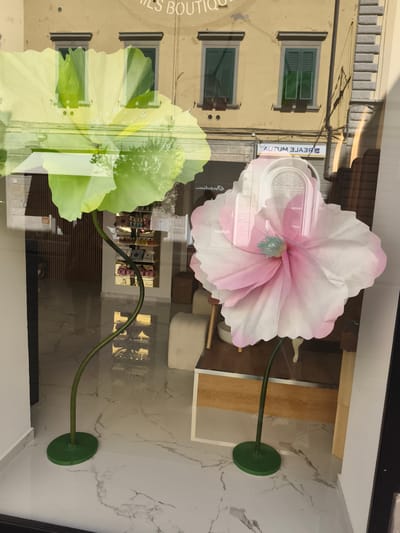 The image shows a street-level storefront window in Livorno, Italy. The window displays two large, artificial flowers. The flower on the left is pale green and the flower on the right is a gradient of pink and white. Both flowers have green stems and bases. Behind the flowers, the interior of the shop is visible, including shelves stocked with various items, likely perfumes or cosmetics, and some furniture. The reflection in the window shows a building with yellow walls and green shutters. A sign on the building reads "AUTUM BJASЯ". The floor inside the shop is tiled with a marble pattern.