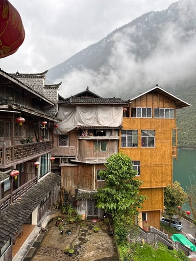 This image depicts a mountainous, riverside architectural scene in Gongtan, China, under an overcast sky. The foreground features a large, traditional multi-story wooden building with dark tiled roofs and ornate wooden railings on balconies, from which several red decorative lanterns are suspended. Directly across a stone-pathed courtyard, a cluster of older wooden buildings with dark tiled roofs descends the hillside. One central structure is partially covered by a white tarpaulin. An exterior air conditioning unit is visible on one of these buildings. To the right, a multi-story building with bright, light-brown wood paneling and numerous modern white-framed windows stands prominently, extending towards a tranquil body of turquoise-green water visible on the far right. This building also features a dark tiled roof with upturned eaves and a small wooden balcony overlooking the water. A dense green tree stands in the mid-ground, partially obscuring the lower levels of the wooden structures. Below the buildings, a paved road runs alongside the water, where a light-colored SUV is parked. Stone steps lead down to the road. The background is dominated by a large, mist-shrouded mountain range under a grey sky, creating a sense of depth and scale. No individuals are visible.