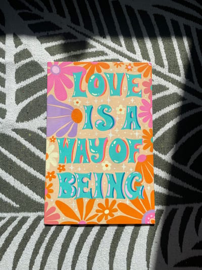 The image shows a painting with the words "LOVE IS A WAY OF BEING" written in colorful, retro-style lettering. The painting is adorned with stylized flowers in shades of pink, orange, purple, and cream. The background of the painting is a light peach color. The painting is resting on a rug with a black and white striped pattern. Sunlight is casting shadows across the rug and the painting, creating a dappled effect. The exact location in the city of Kopeysk, Russia, is not discernible from the image.
