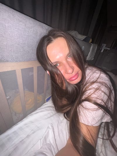 The image features a young person, likely female, with long, straight dark brown hair and greenish eyes, lying on a bed and looking directly at the camera. She has a slight, relaxed smile. She is wearing a white t-shirt.
The setting is an indoor bedroom, characterized by soft, possibly dim, lighting. The bed is covered with white sheets that display a subtle woven or striped pattern. To the person's left, a grey upholstered surface, likely a headboard or side of the bed, is visible. To her right, a light-colored, fine-mesh barrier, possibly a bed rail or part of a cot, extends across the mid-ground.
A notable object in the upper left background is a dark-colored item resembling an assault rifle, which appears to be mounted or leaning against a dark wall or piece of furniture. No visible text is discernible on the person's clothing or the immediate bed linens. The overall impression is one of a casual, private moment.