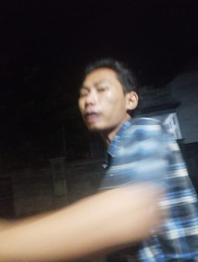 The image captures a person, likely male, in a dimly lit or nighttime setting, characterized by significant motion blur. The subject's head and upper torso are visible against a dark, indistinct background. He appears to be of Asian descent with short, dark hair. His eyes are partially closed or squinting, and his mouth is slightly open, suggesting he might be speaking, reacting, or caught mid-expression. He is wearing a blue and white plaid shirt, which, like the rest of the subject, exhibits noticeable blur, indicating movement during the exposure. A blurry, light-skinned arm or hand extends into the foreground from the lower left. Faint outlines of structures can be vaguely seen in the dark background on the right. The overall impression is one of movement or a brief, candid moment captured in low light. No visible text is present in the image. The location context for the image is Kecamatan Jangkar, Indonesia.