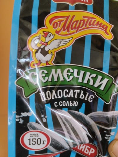 The image shows a close-up of a food product packaging, specifically a bag of sunflower seeds. The packaging has a black background with vertical blue stripes. Dominating the top of the bag is a yellow banner with red lettering that reads "от Мартина". Below this banner, a cartoon chicken wearing a red bow tie and vest is depicted.
The main product name, written in white, bold, stylized letters within a green curved banner, reads "СЕМЕЧКИ". Beneath this, in white, slightly smaller font, it says "ПОЛОСАТЫЕ", indicating striped sunflower seeds. Further down, in smaller white text, it says "С СОЛЬЮ", meaning "with salt".
In the lower left corner, a circular red and white label displays "МАССА НЕТТО:" (NET WEIGHT:) above the number "150г" (150 grams). The packaging features images of sunflower seeds, some of which are partially obscured by the plastic wrap's reflections. A person's thumb is visible in the lower right corner, suggesting the product is being held.
The overall impression is of a commercially produced food item, likely a snack. The colors are bright and the cartoon image adds a playful element. The text is in Russian, and the inclusion of "Павловское, Russia" in the prompt suggests this is the likely origin or point of sale for the product. There are no indications of the time of day, weather, or specific emotions conveyed, other than the general cheerful marketing of a snack product.