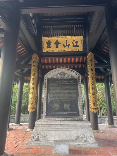 This is a photograph of a monument located in the city of Phong Châu, Vietnam. The monument is situated under a dark wooden pavilion with a red-tiled roof. A golden plaque with black calligraphy is mounted on the pavilion above a large stone tablet. Flanking the tablet are two ornate golden pillars with more calligraphy. The stone tablet itself is weathered and inscribed with Vietnamese text. The ground in front of the monument is paved with red bricks. The background shows lush green trees and foliage, indicating an outdoor setting. The image is well-lit, suggesting it was taken during the daytime. There are no people visible in the photograph.