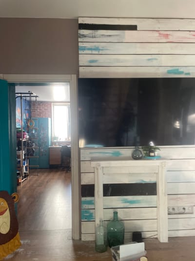 The image depicts an interior scene, likely a living room, viewed from a doorway. A wall with a rustic, horizontally paneled design, painted in white with streaks of black, red, and blue, dominates the right side of the frame. A large, dark television screen is mounted on this wall. Below the television, a white mantelpiece-like structure also features the paneled design. Two green glass bottles and a small potted plant are arranged on this mantelpiece.

To the left of the frame, a doorway opens into another room. Through this doorway, a wooden floor and a wall made of exposed brick are visible. In this adjacent room, there is a tall, teal locker, a wooden desk with a chair, and a window letting in bright sunlight. There is also a shelf unit with various items, including what appears to be a climbing rope and rings. A decorative lion cutout is partially visible on the left edge of the frame, near the doorway. The overall impression is of a lived-in, eclectic space.

The location is identified asМухавецкі сельскі Савет, Belarus.