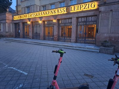 The image shows the exterior of a building in Leipzig, Germany, with the illuminated text "ALLGEMEINE ORTSKRANKENKASSE LEIPZIG" above a large entrance with ornate metal gates. In the foreground, two pink electric scooters are parked on a paved area. One scooter, in the immediate foreground, is a "VOI" brand scooter, identifiable by the logo on its stem and the green light on its handlebars. The other scooter is partially visible on the right. The building appears to be an administrative or institutional building, possibly a health insurance office, given the text. The time of day seems to be late afternoon or early evening, as indicated by the lighting. The environment is urban, with the building's facade and the paved plaza. There are no people visible in the image.