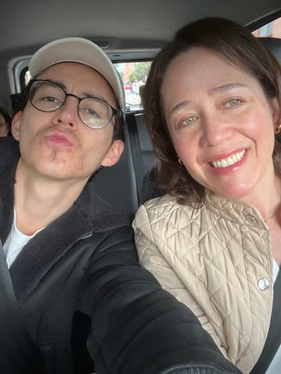 The image depicts three individuals inside a passenger vehicle during daytime. In the foreground, a young adult male is positioned on the left, wearing a light-colored baseball cap, black-rimmed glasses, and a black jacket with a dark, fuzzy collar over a white t-shirt. He has sparse facial hair (mustache and chin tuft) and multiple red blemishes on his cheeks and chin. His lips are pursed in a pouting expression. To his right, an adult female with brown hair and green-brown eyes is smiling broadly, revealing her upper teeth. She is wearing a light beige quilted jacket and small gold hoop earrings. Both individuals have engaged seatbelts crossing their chests. A third individual, partially obscured, is visible in the rear on the far left, showing part of their face and dark hair. The vehicle's interior features black seat upholstery. Through the vehicle window in the background, an urban street scene is visible. This scene features multi-story buildings constructed primarily of reddish-brown brick, a paved road with white lane markings, green foliage (trees), and distant pedestrians. The distinctive red-brown brick architecture is characteristic of urban areas within Bogota, Capital District, Colombia. The overall scene suggests a casual social interaction, possibly a selfie, taken while traveling or parked in the city.
