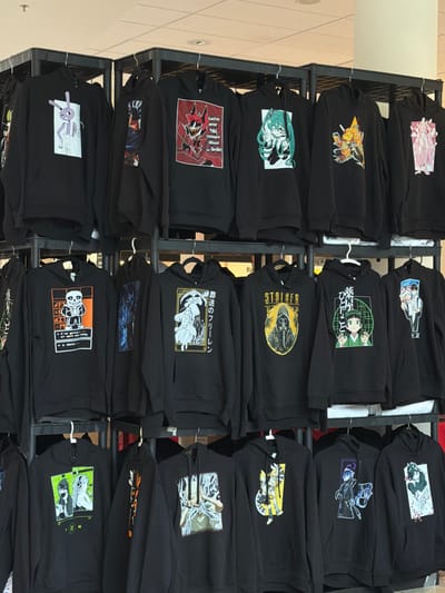This media file shows a retail display of black hoodies featuring anime-style graphics. The hoodies are neatly folded and arranged on black metal shelves, likely in a clothing store. The graphics on the hoodies depict various characters and scenes, common in anime and manga. Some of the visible text includes Japanese characters and phrases like "You're Never Dressed Without a Smile" and "I've gotten a lot of work done today." There is also text associated with "STALKER" and "PELET." The lighting appears to be artificial, suggesting an indoor environment. The overall impression is that of a merchandise display for fans of anime and related pop culture. The scene is static, with no people actively interacting with the display, creating a sense of organized retail presentation.