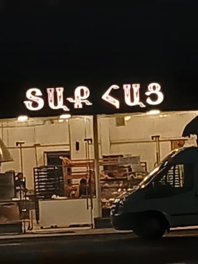 A nighttime exterior shot shows a brightly lit sign with white Armenian letters against a dark background. The sign reads "SULUS". Below the sign, a building's facade is visible, illuminated by yellow lights. The building appears to be a commercial establishment, possibly a bakery or food service business, with shelves stocked with items and visible ovens or preparation areas through open doorways. A dark-colored van is parked in the foreground on the right side of the image, partially obscuring the building. The van's headlights are not on. The street in front of the building is dark and appears to be paved. The overall scene suggests an active business operating late at night in the city of Vagharshapat, Armenia.