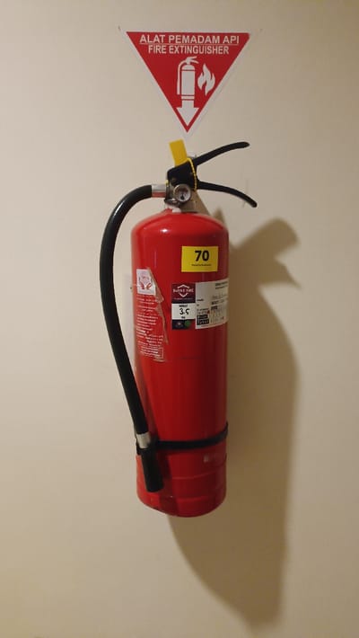 A red fire extinguisher is mounted on a plain, light-colored wall in Batulawang, Indonesia. Above the extinguisher, a red triangular sign with a white border displays the text "ALAT PEMADAM API" and "FIRE EXTINGUISHER" in white lettering, accompanied by a white graphic depicting a fire extinguisher, a flame, and a downward arrow.
The fire extinguisher itself is red, featuring a black curved hose with a black nozzle attached to its side. At the top, a black handle assembly includes a silver-colored pressure gauge and a yellow plastic safety pin/seal. Affixed to the cylindrical body are two primary labels: a yellow rectangular label prominently displaying "70" with smaller text "Hotel Lie Enn" below, and a larger white label with a red logo. This white label details "BADIKE FIRE", "PT. BADIKE FIRE INDONESIA", and specifies a "BERAT: 3.5 KG" (weight: 3.5 kg). Other partially visible text includes "TABUNG 2024". The extinguisher is secured to the wall via a black mounting strap positioned around its lower half. Lighting from the left casts a distinct shadow of the extinguisher and its components onto the wall to the right. No people or other objects are present in the frame.