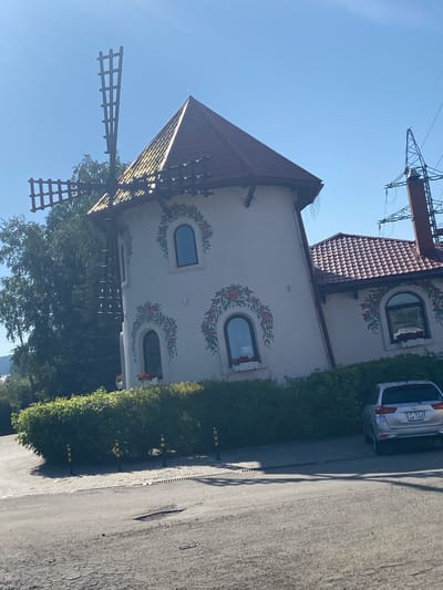 A white, multi-story building with a conical, red-tiled roof and a windmill structure attached to its side is visible under a clear blue sky. The building has arched windows decorated with floral motifs. A section of a larger building with a similar red-tiled roof is attached to the main structure. A silver SUV is parked to the right of the main building, in front of a dense green hedge. Several black and yellow bollards are placed along the edge of the hedge. The foreground shows a paved area with some debris and tire marks. To the right of the building, a tall red brick and metal electricity pylon is present. The scene appears to be in a suburban or semi-rural area.