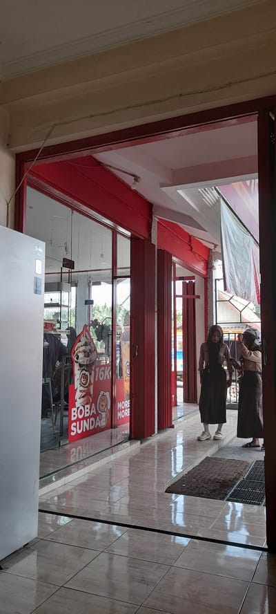 The image depicts the entrance to a "Momoyo" boba tea shop, likely located in Crewek, Indonesia. A large, white refrigerator is visible on the left. The shop features a red-painted facade with large glass windows. A prominent advertisement on the window displays a large ice cream sundae with the text "BOBA SUNDAE" and "16K," indicating a price. Two young women in hijabs are standing in the entryway. One is wearing a light brown uniform and a dark hijab, looking towards the other woman and holding a phone. The second woman is wearing a similar uniform and hijab, with a white helmet on her head. She appears to be adjusting the first woman's hijab or helmet. The floor is tiled and reflects the overhead lighting. The overall scene suggests a retail environment, possibly a franchise shop, with a focus on desserts and beverages.