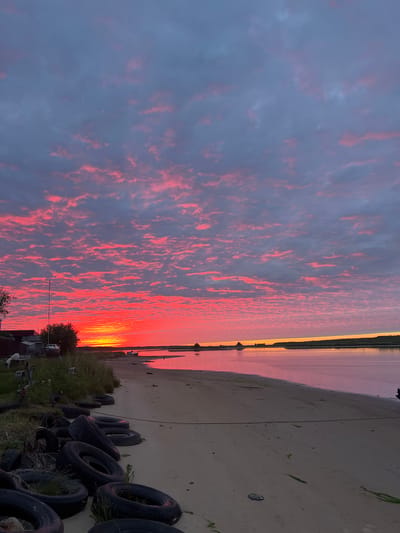 The image captures a vibrant sunset over a body of water near the city of Severodvinsk, Russia. The sky is filled with clouds illuminated by intense shades of red, orange, and pink, creating a dramatic effect. The water reflects these colors, appearing as a soft pinkish hue. The foreground features a sandy beach with numerous discarded tires scattered across it. Some tires are partially overgrown with grass and small plants. Behind the beach, there is a strip of vegetation, including grass and trees. In the background, a few buildings and a sailboat are visible along the shoreline. The overall scene depicts a natural landscape at dusk, with evidence of human activity in the form of the discarded tires.
