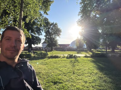 The image is a selfie taken outdoors, likely on a sunny day in Târgu Mureș, Romania. A person's face, specifically the right side, is visible in the upper right portion of the frame. The background includes a green lawn, trees, and a building. Sunlight is present. The person is wearing a dark-colored jacket. The image is taken at an unusual angle, rotated roughly 180 degrees. In the background, a building with a glass dome-like structure is seen, potentially indicative of the location in Târgu Mureș.
