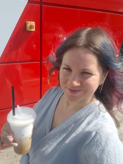 A woman is standing outdoors, holding a plastic cup with a beverage and a straw. She is positioned in front of a red and white building. The woman has brown hair with blue and pink highlights. She is wearing a light blue top. A yellow box with a red button is on the red building. The image was taken in Sholokhovo, Russia.