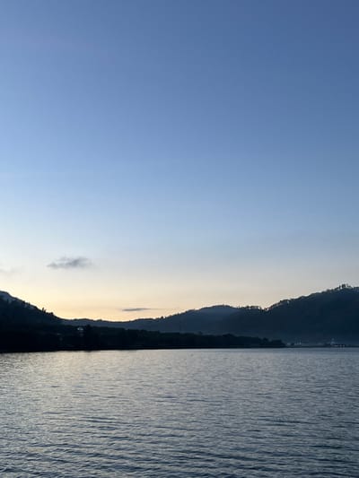 The image depicts a serene body of water with mountains in the background. The sky is a clear gradient of blue, transitioning to a lighter hue near the horizon where a faint glow suggests either sunrise or sunset. A small, dark cloud is visible in the upper left quadrant of the sky.
The water surface is textured with gentle ripples, reflecting the soft light of the sky. Along the horizon, a chain of dark, tree-lined mountains forms the backdrop. The silhouettes of the mountains are softened by a light haze or mist, particularly in the distance, giving the scene a tranquil and somewhat ethereal quality. On the lower slopes of the mountains, there are indications of vegetation, with distinct tree lines visible in places.
In the mid-ground, on the shore between the water and the mountains, there are hints of human settlement. Lights can be seen in the distance, suggesting buildings or structures. On the far right, a building with a dome-like structure is visible, possibly a mosque or a similar religious building, which aligns with the location in Merodot, Indonesia, a region known for its Islamic heritage. The overall impression is one of a peaceful natural landscape with subtle signs of habitation.