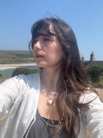A young woman with long, dark, wavy hair and bangs is shown in a close-up, outdoor selfie. She is looking to her right, with her head tilted slightly upwards. She is wearing a white, open-fronted shirt over a dark gray tank top. A silver necklace with a white, heart-shaped pendant hangs around her neck.
The background reveals a coastal landscape under a clear blue sky. A pebbled beach meets a body of calm, turquoise water, with several small figures visible on the beach. Rolling green hills rise behind the beach. In the middle ground, a red-tiled roof with two prominent brick chimneys is visible, suggesting a building situated on the hillside. The environment appears sunny and warm, typical of a seaside location. The image was taken in Seaford, United Kingdom.