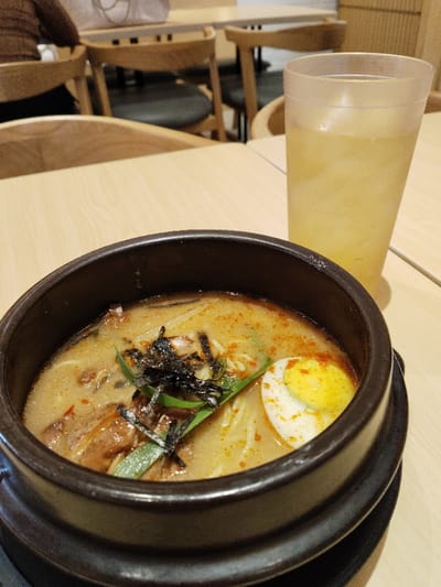 A bowl of ramen with a soft-boiled egg, seaweed, and green onions sits on a wooden table. A glass of iced tea sits next to the bowl. The table is in a restaurant setting, with blurred chairs and tables in the background. The ramen is situated on a table in a restaurant in Telukjambe, Indonesia, at coordinates -6.301, 107.278. The ramen is in a dark ceramic bowl, with a brown broth and noodles. The egg is partially submerged in the broth, and the seaweed and onions are placed on top. The tea is light in color and slightly cloudy.