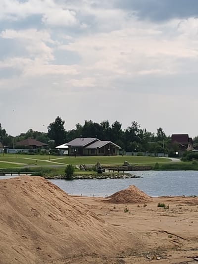 The image depicts a residential area near a body of water, likely a lake or river, in Vasil'ki, Russia. The foreground is dominated by a large pile of sand, suggesting construction or landscaping activities. Behind the sandpile, a body of blue water stretches across the midground, with a few small birds visible on the shore.
In the background, a manicured green lawn slopes down to the water, dotted with streetlights and pathways. Several houses with dark-pitched roofs are visible, some appearing modern and single-story, while others have more traditional architectural features. One house, in particular, has a prominent white awning extending from its facade. Dense green trees form a backdrop to the houses, suggesting a wooded or park-like setting. The sky above is overcast with a mix of light and dark clouds. The overall scene conveys a sense of suburban development with natural elements.