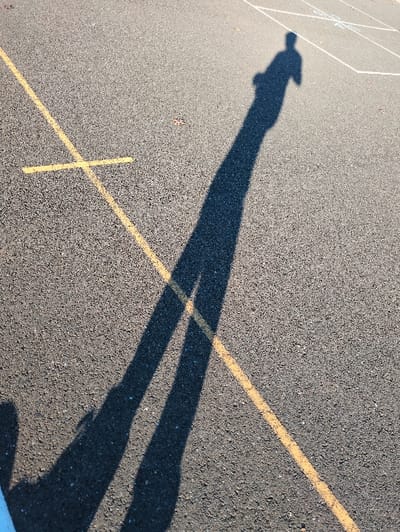 A stark black shadow of a figure stretches across asphalt, divided into two legs near the end. A yellow line bisects the shadow and extends into a perpendicular line, forming a cross shape. A faded hopscotch grid is visible in the upper left corner. The asphalt has a textured, grainy appearance.