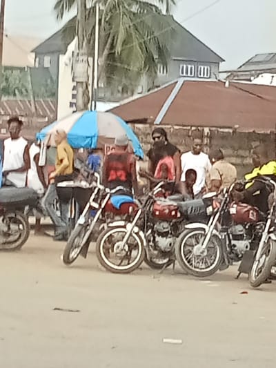 The image displays an unpaved roadside scene in Orie Uratta, Nigeria. In the foreground and midground, a grouping of at least six motorcycles, predominantly red and black models, are parked or idling. Several adult males are positioned around and on these vehicles. A man, viewed from the rear, wears a red sleeveless shirt with white numbering (possibly "22") and is situated on a motorcycle with a blue fabric draped over its seat. Adjacent to him, a man with dark skin, wearing a black sleeveless top and sunglasses, with visible dreadlocks or braids, stands near a red motorcycle. A child in a red shirt is partially visible in front of this man. Further right, an individual in a white short-sleeved shirt faces the viewer. To the left, a man in a yellow long-sleeved shirt stands near a partially open blue and white striped market umbrella. On the far right, an individual in a bright yellow top is seated on a motorcycle. The background features a large palm tree, which partially obscures a two-story building with a dark grey roof and white walls. Additional structures with reddish-brown roofs are visible below and to the right of the main building. The environment indicates a local gathering point or motorcycle taxi (okada) stand.
