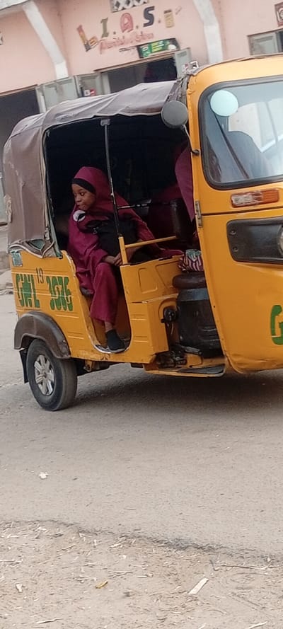 A young girl, wearing a deep maroon or pink hijab and matching long dress, sits inside a yellow three-wheeled auto-rickshaw, commonly known as a 'keke napep' in Nigeria. She is positioned towards the left side of the vehicle, looking out with a neutral expression. Another individual, similarly dressed, is partially visible deeper within the rickshaw. The vehicle itself is painted bright yellow with a dark brown soft top. On its side, faded green numbers and letters are visible, reading "19 GML 3676". A partial green letter "G" is also seen on the front-right panel of the rickshaw.

The auto-rickshaw is parked or moving slowly on a dusty, unpaved road. The ground is light brown with scattered debris and small pebbles. In the background, there is a light-colored building, likely made of concrete, with a prominent sign featuring large, stylized red and brown letters spelling "I.A.S.". Below this, smaller, less distinct text appears to read "Furniture Home". To the right of "I.A.S.", another sign, possibly green, is visible, though its text is not clearly discernible.

The scene takes place during the daytime under bright, natural light, suggesting clear weather. The overall impression is a common street scene in a Nigerian town or city, typical of the location context of Kabuga.