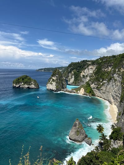 The image shows an aerial view of Kelingking Beach in Pejukutan, Nusa Penida, Indonesia. A large, jagged rock formation covered in vegetation juts out into the turquoise ocean. At the base of the formation is a white sand beach where waves are crashing. Smaller, similar rock formations are also visible in the surrounding water. Above the rock formations and ocean is a blue sky with scattered white clouds. A thin cable stretches across the sky in the upper left corner of the image.