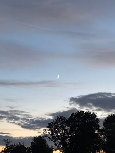 This is a photograph taken at dusk, likely in a rural area given the prominent silhouettes of trees against the sky. The sky is a gradient of blues and grays, with wispy clouds suggesting the end of daylight. In the upper portion of the frame, a slender crescent moon is visible, accompanied by a single bright star. The lower part of the image is dominated by the dark, leafy silhouettes of trees, with the faintest hint of orange light on the horizon below them, indicative of a sunset. The scene is tranquil and atmospheric, capturing a moment of celestial beauty as night approaches. The location in Ганчарскі сельскі Савет, Belarus, suggests a landscape characterized by open spaces and natural vegetation, consistent with the image's content. There are no people or man-made structures clearly visible, emphasizing the natural setting.