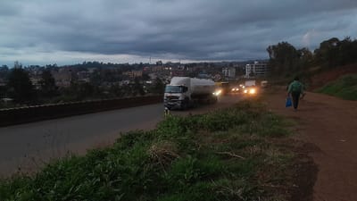 The image shows a view from a hill overlooking a highway in Gitaru, Kenya. It is early evening and the sky is overcast with a few streaks of blue. In the foreground, a person walks along a dirt road, carrying a blue shopping bag. In the distance, a large truck is driving along the highway. The truck is silver and has a large tank on the back. Behind the truck, there are several other cars and vehicles driving in the same direction. The road is bordered by a concrete wall and a grassy area. The city of Gitaru is visible in the background, with many buildings and houses. The environment is rural and there are trees and bushes along the sides of the road. The image captures a quiet moment on a road in Gitaru.