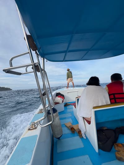 From the edge of a moving boat, a man stands at the edge wearing a hat, light shirt, and tan shorts. Inside the boat, a child wears a red life vest, and a person in a white towel sits near a pair of tan flippers and a black backpack. The blue sky is cloudy. The boat has a blue roof with a silver rail and metal brackets. Water rushes past the boat.