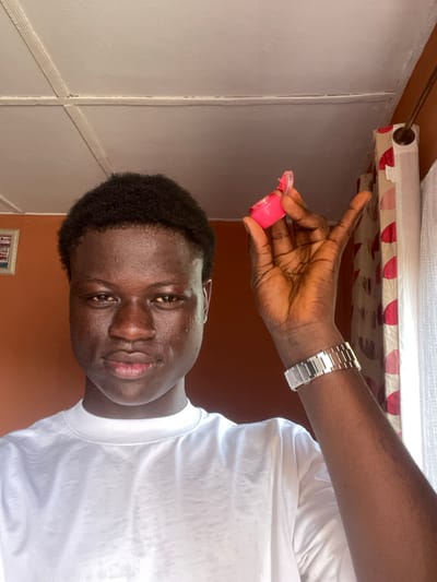An indoor, waist-up portrait captures a young adult male with dark skin and short, dark, curly hair, facing directly forward. He wears a plain white crew-neck t-shirt and a silver-toned metal watch on his left wrist. His right hand is raised, holding an open, small, circular, bright pink container, likely a lip balm, above his head to the right. The background features an orange-brown wall to the left, with a partially visible framed picture. To the right, a window is covered by a sheer white curtain, behind which a patterned curtain with red and white circular motifs hangs from a brown rod. The ceiling is white with visible structural lines. The scene is located in Mosafejaw, Nigeria.