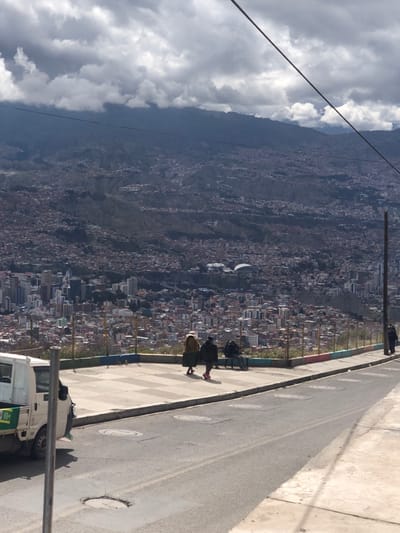 The image displays an expansive, elevated view of a vast city sprawling across a mountainous or hilly landscape, captured from what appears to be a roadside overlook. The scene is illuminated by daylight, though the sky is largely covered with dramatic, dense grey and white clouds, suggesting a cloudy day with intermittent brightness.

In the background, the city extends far into the distance, with countless buildings, primarily residential, densely packed onto the slopes and valleys of the terrain. A prominent feature within the urban sprawl is a large, white, dome-shaped structure, standing out among the other buildings. The distant horizon is defined by rugged, dark mountains that merge with the overcast sky.

In the midground, running along the right side of the image, is a paved road or a wide pedestrian path. A metal fence or railing separates this elevated area from the steep drop-off overlooking the city. The base of this railing is adorned with colorful painted sections. Several people are visible near the fence. Two individuals, dressed in what appears to be traditional attire including skirts and shawls, are standing or walking along the path, seemingly taking in the view. A utility pole stands upright near the center-right of the frame, extending above the cityscape.

In the foreground, a white utility truck is parked on the side of the paved road in the lower right corner. The truck has a green and white logo or sticker on its side, though specific details or text are not clearly legible. The road itself features several circular covers embedded in its surface. A thin wire or cable stretches diagonally across the upper portion of the sky, possibly part of a power line or a cable car system.