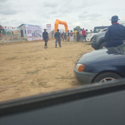 The image depicts an outdoor scene in Wuro Ngangandi, Nigeria, with a crowd gathered for a ground-breaking and commissioning event. In the foreground, a dark blue car is visible, partially obscuring the view. Further back, several individuals in uniforms, likely security personnel, are present. One uniformed individual, wearing a dark uniform and a red cap, walks towards the right of the frame, holding a rifle. Another uniformed individual in camouflage is also visible, facing away from the camera.

Behind the security personnel, a large orange inflatable arch stands, possibly marking an entrance or a ceremonial point. A gathering of people, dressed in casual clothing, is positioned behind the arch, with some looking towards the event. To the left of the arch, a small temporary structure with a pitched roof is situated, and a banner on its facade announces "GROUND BREAKING AND COMMISSIONING" and mentions "IN LINE WITH PRESIDENT BOLA AHMED TINUBU RENEWED HOPE AGENDA". Several other banners and flags are also displayed in the background.

The environment is an open, dirt-covered area, indicative of a construction site or an outdoor event venue. The sky is overcast with clouds. In the far background, there are some trees and structures. The overall scene suggests a public ceremony or event taking place.