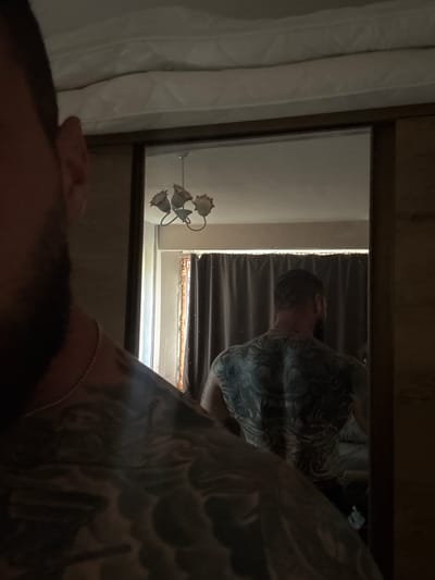 In a dimly lit room, a man with extensive back tattoos is reflected in a mirror. The man is facing away from the camera, and his head and shoulders are partially visible in the foreground. The mirror shows his back, covered in intricate, dark-inked designs that appear to be of mythical creatures or oriental motifs. He is wearing a thin necklace. Behind the man in the reflection, dark curtains are drawn across a window, and a light fixture with multiple flower-shaped lamps hangs from the ceiling. The room appears to be a bedroom, with a white mattress visible at the top of the frame, possibly on a bed or bunk. The overall environment suggests a domestic setting. The location is identified as Radomir, Bulgaria.
