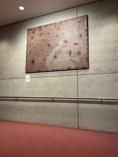 An abstract or patterned artwork is prominently displayed on a textured, light gray wall in an indoor setting, likely a gallery or museum in South Brisbane, Australia. The artwork itself is a vertical rectangular piece, rich in detail. It features a multitude of small, irregular light-colored dots or strokes set against a darker, possibly maroon or reddish-brown background. Interspersed within this intricate texture are larger, more defined circular and oval shapes, each characterized by a vivid red center encompassed by a gray or darker outline.

The gray wall, where the artwork is hung, appears to be constructed from large rectangular panels, discernible by the subtle grout lines. Illumination comes from a spotlight positioned above and to the left, creating a slight glare on the artwork's surface and casting a soft shadow beneath it. To the left, a portion of a lighter-colored, angled wall is visible, featuring a recessed circular light fixture.

To the right of the artwork, a thin, vertical metal pole runs along the gray wall, fitted with small hooks, suggesting it's part of a flexible display system. Further to the right, a solid pink or reddish-orange wall provides a striking color contrast to the neutral tones of the main display area. A small, white informational card is attached to the wall below the artwork, but any text on it is too small and blurry to be deciphered. The overall scene suggests a contemporary art exhibit.