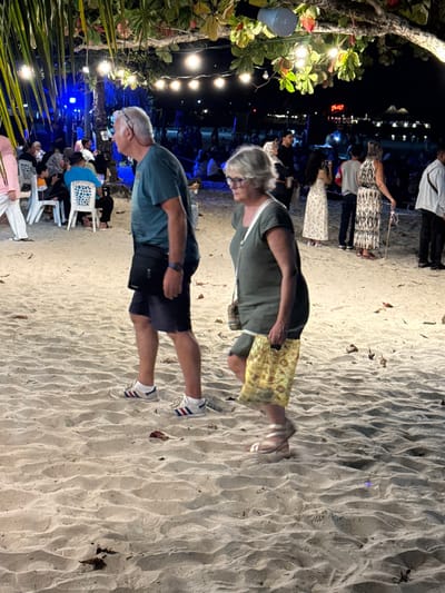 The image depicts a nighttime scene on Cenang Beach, Malaysia. In the foreground, two elderly individuals are walking left across the sandy terrain. The man, with short grey hair, wears a teal short-sleeved shirt, dark shorts, white athletic shoes with red and blue stripes, and a black messenger bag slung across his body. The woman, with grey hair and glasses, wears a dark olive-green short-sleeved shirt, a patterned yellow reusable shopping bag in her right hand, and light-colored sandals. The sand beneath them shows ripples and scattered small dark objects, possibly leaves or debris.

In the midground and background, numerous other people are present. To the left, a group is seated at white plastic tables with chairs under a blue light source, suggesting an eating or gathering area. Further back, more people are seated directly on the sand. Overhead, string lights with incandescent bulbs are suspended from tree branches, providing illumination for the scene. A solitary light fixture with a blue shade hangs from a branch directly above the main subjects. In the far background, additional lights from buildings or other structures are visible against the dark sky, including a red illuminated sign that reads "THE V". On the right side of the midground, several individuals are standing, including a woman in a long patterned dress holding what appears to be a walking stick or cane, and another woman in a light-colored long dress holding a phone, possibly capturing a photograph. The overall atmosphere is that of a lively evening social gathering on the beach.