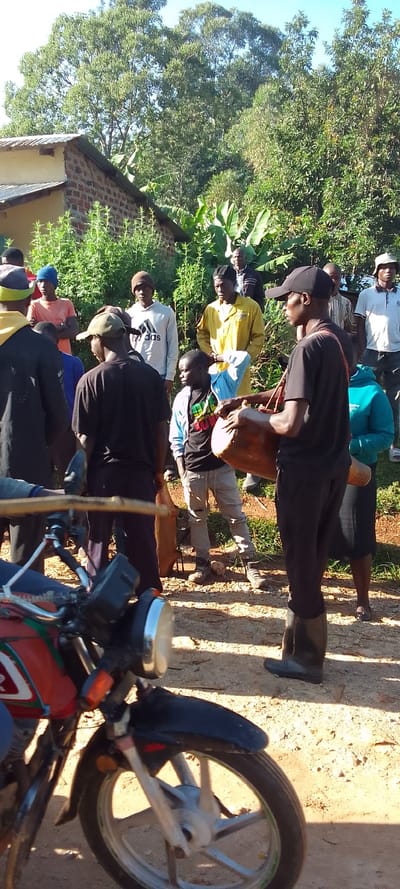 The image depicts a group of people gathered in a rural setting, likely near a small village or settlement, in Isukha ICHINA, Kenya, located at coordinates 0.352 latitude and 34.782 longitude. There is a motorcycle parked in the foreground. It is a partially sunny day, and the ground is dusty and brown, likely dry from a lack of rain. The group is primarily composed of men, with a few women and children present. They are wearing a mix of casual and traditional clothing. Some individuals are standing, while others are sitting on a wooden log or bench. In the center of the scene, a man is holding and beating a large drum. Other individuals are engaged in conversation, or watching the performance. There is a small house or building visible in the background, and the scene is surrounded by vegetation. This appears to be an informal community gathering or celebration. The scene captures a moment of communal activity and interaction within the Isukha ICHINA community.