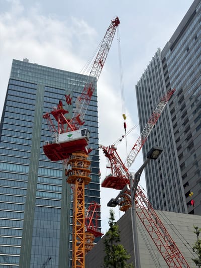 This is a photograph taken in Chiyoda, Japan, of a construction site. The image features two large red and white tower cranes, a prominent orange construction tower, and two modern high-rise buildings. One building, to the left, is clad in glass and features a grid-like pattern of windows. The other building, to the right, is made of gray material and also has a regular pattern of windows. The sky is overcast with some clouds. The cranes are in various stages of operation, with some equipment visible. A streetlight and some green trees are also present in the foreground.