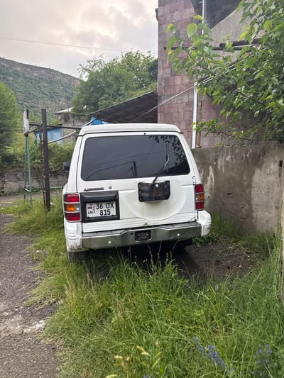 In the city of Tumanyan, Armenia, a white Mitsubishi Pajero SUV is parked on a dirt path adjacent to a concrete wall. The vehicle's rear is facing the camera, displaying an Armenian license plate with the number 36 OX 815. A spare tire mount is attached to the rear door. Overgrown green grass and some small blue flowers surround the lower portion of the vehicle and the path. Behind the SUV, a partially visible building with a stone facade and a corrugated metal roof can be seen. Further in the background, a steep, tree-covered mountainside rises under an overcast sky. A metal fence structure is also present to the left of the vehicle.