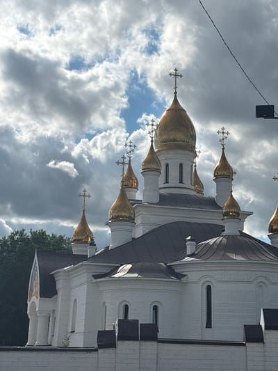 The image is a low-angle shot of white and gold-domed religious architecture against a cloudy sky. The domes are ornate and topped with multi-tiered crosses. The building's facade is white brick with arched windows and decorative moldings. Dark gray roofs with curved lines are visible, contrasting with the white walls. The scene is likely a church or cathedral. The sky is filled with dramatic, textured clouds, with patches of blue sky visible. Sunlight reflects off the golden domes, suggesting it is daytime, likely late afternoon given the lighting and cloud cover. There is a dark, rectangular object suspended by wires in the upper left corner, which could be a lighting fixture or part of an electrical system. The overall mood is somewhat somber due to the heavy clouds, yet the golden domes add a touch of grandeur and hope. There are no people visible, and no text is discernible. The context provided is Orsk, Russia, which helps place this as a religious site in that region.