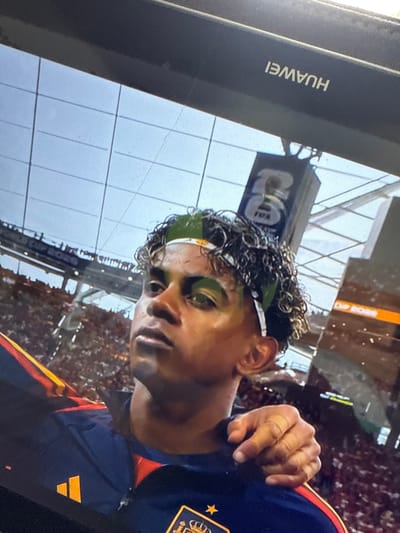 The image shows a close-up of a young man with curly dark hair and a headband. He is wearing a dark blue sports jersey with red and yellow accents. There is an orange Adidas logo on his left shoulder. His right arm is draped over his own shoulder, and his hand is visible in the foreground, with what appears to be tape on his fingers. In the background, there is a stadium filled with spectators. A large screen or scoreboard is visible above the crowd, with the FIFA logo on it. The brand name "HUAWEI" is visible on a dark frame at the top of the image, suggesting the photo was taken of a screen displaying a game. There is no information to suggest this image is located in Almada, Portugal.
