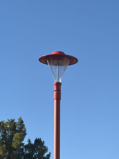 A red street lamp with a clear glass cover stands against a clear blue sky. The lamp has a wide, flat red top and a conical glass section that tapers down to a narrower red tube. The lower part of the lamp post is also red. To the left of the lamp post, the upper branches of a green coniferous tree are visible. The image appears to be taken in Schwedt/Oder, Germany, based on the provided location context.