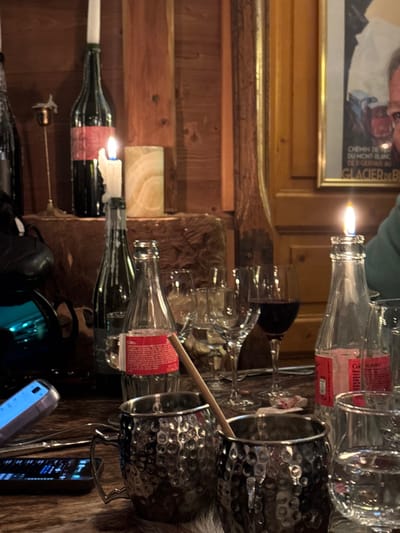 This image captures a cozy, rustic indoor scene, likely in a restaurant or bar in Val-d'Isère, France, during the evening. The main subjects are an assortment of glassware and bottles arranged on a surface, possibly a wooden bar or table, partially covered by a furry hide.
In the foreground and midground, several empty clear glass bottles are visible, one with a red label. Two bottles serve as makeshift candle holders, with lit candles emanating a warm glow, and melted wax dripping down a dark green wine bottle. A collection of empty wine glasses and tumblers are stacked and placed nearby. To the right, two metallic, hammered-texture mugs are seen within a metal caddy, one containing a wooden stirring stick. A smartphone displaying information lies on the right side of the frame, alongside a dark, reflective object.
The background features warm, rustic wooden paneling, typical of an alpine chalet. A framed picture hangs on the wall, showing a close-up of a man's smiling face. Visible text on the picture includes "CHEMIN DE FER DU MONT BLANC DES SERVOZ AU" and "GLACIER DE BLANCS", clearly indicating a connection to the Mont Blanc region in the French Alps, reinforcing the location context.
The scene suggests a relaxed gathering or dining experience, highlighted by the use of candlelight for ambiance. The lighting indicates it is nighttime or a dimly lit interior. No people are actively present in the immediate scene, only their traces in the form of used glassware. The overall atmosphere is intimate and comfortable, characteristic of a mountain resort establishment.