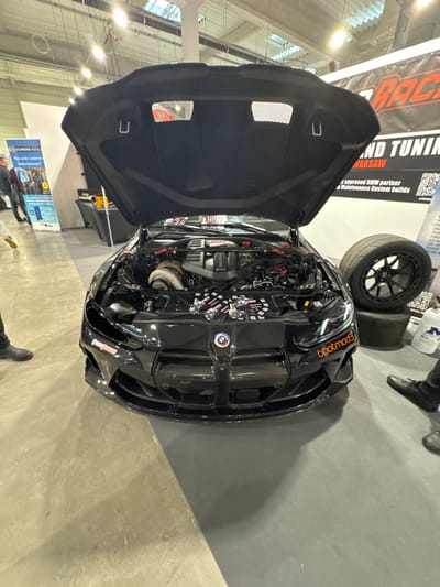 A modified black BMW M-series performance car is centrally displayed indoors at an exhibition, identified as likely being at the Ptak Warsaw Expo in Wolica, Poland. The vehicle's hood is open, showcasing a heavily customized engine bay with a prominent turbocharger on the left, a BMW M-branded intake manifold, and red-accented structural components. Several small, assorted items are arranged on a support bar across the front of the engine bay. The car features a carbon fiber front bumper and grille, with an orange "bootmod3" decal on the front bumper's right side.

To the right of the vehicle, a large black aftermarket wheel fitted with a slick racing tire is propped against two smaller, worn tires. A white plastic jug with a blue label is positioned on the floor adjacent to the tires. The feet of a person wearing black dress shoes are partially visible on the far right.

In the background, a blue and white "BOSCHMA.AUTO" banner stands on the left, near which a man in a dark jacket and blue jeans is partially visible. On the right, a large black banner with red and white lettering reads "GRAND TUNING WARSAW" and details "An approved BMW partner, Maintenance, Custom builds," with additional smaller banners below. The floor is a light grey, reflective surface, consistent with an exhibition hall environment. Overhead industrial lighting fixtures are visible on the ceiling.